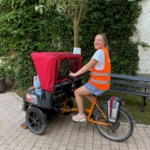 Eine junge Frau mit orangenen Warnweste fährt als Flaniermobil-Pilotin ältere Menschen durch die Stadt.