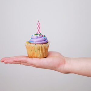 Auf einer Hand liegt ein bunter Cupcake mit einer Geburtstagskerze drauf.