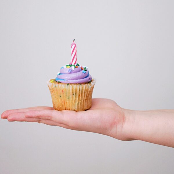 Auf einer Hand liegt ein bunter Cupcake mit einer Geburtstagskerze drauf.