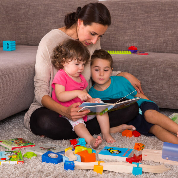 Eine Mutter sitzt vor der Couch auf dem Boden und schaut mit ihren zwei Kindern in ein Bilderbuch. Um die Drei liegen Spielzeug verteilt auf dem Boden.