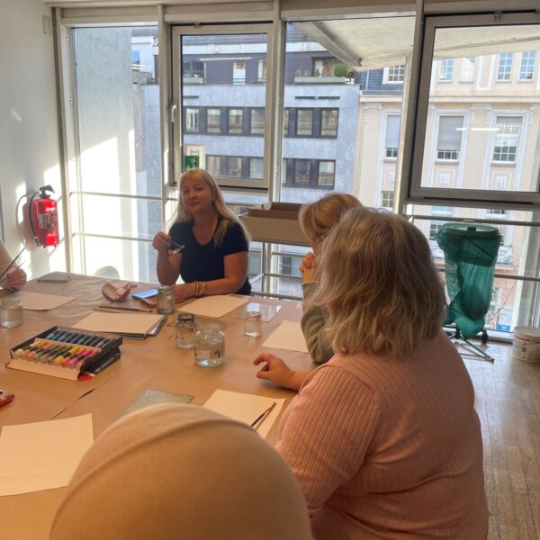 Eine ehrenamtliche Kunsttherapeutin aus der Ukraine sitzt im Rahmen eines interkulturellen Workshops mit Teilnehmerinnen zusammen am Tisch. Vor ihnen liegen Papier und Stifte.