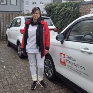 Eine Kollegin in Caritas-Kleidung steht vor einem weißen Wagen mit Caritas-Logo.