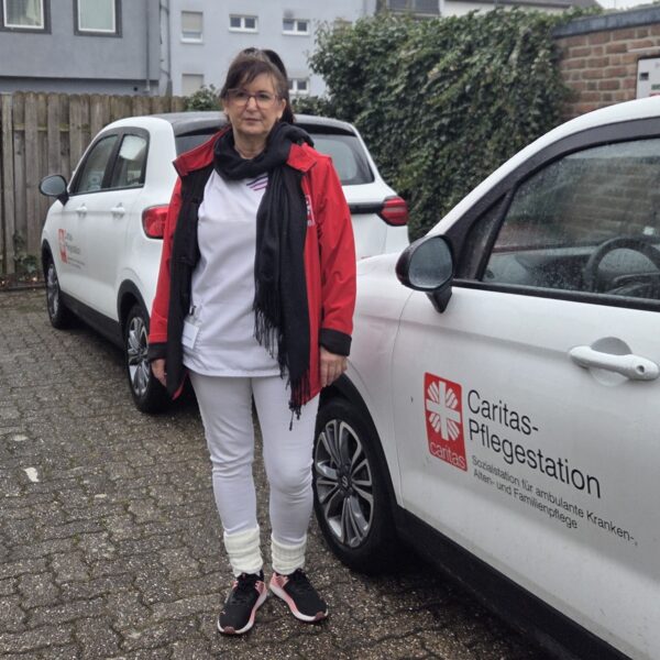 Eine Kollegin in Caritas-Kleidung steht vor einem weißen Wagen mit Caritas-Logo.