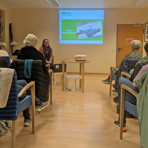 Auf dem Bild ist die Referentin, sitzend vor ihren Powerpointfolien zu sehen sowie einige der Anwesenden sitzend auf Stühlen von hinten.