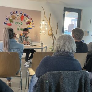 Zwischen den Köpfen der Gäste schaut man auf den Autor, der an einem Tisch sitz und seine Geschichte liest. Über ih, hängt ein Plakat mit der Aufschrift Haus Christophorus.