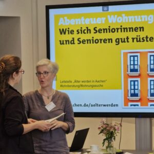 Zwei Frauen unterhalten sich angeregt vor einer PowerPoint-Präsentation zum Thema Seniorenarbeit.