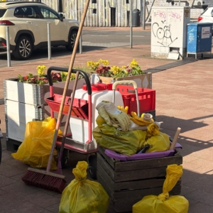 Gut gefüllte gelbe Müllsäcke und ein Besen lehnen an einem Blumen-Hochbeet.