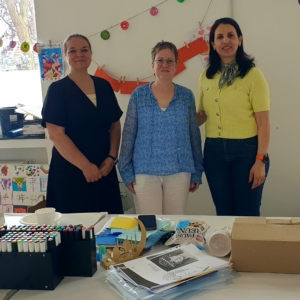 Unsere Kollegin Géraldine Wronski, Caroline Strasser von der Beratungssstelle und die ehrenamtliche Kollegin Nibal Farhat stehen vor einem Tisch und lächeln in die Kamera.
