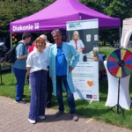 Eine Mitarbeiterin und ein Ehrenamtler posieren vor dem Infostand, Webebanner und dem Glücksrad.
