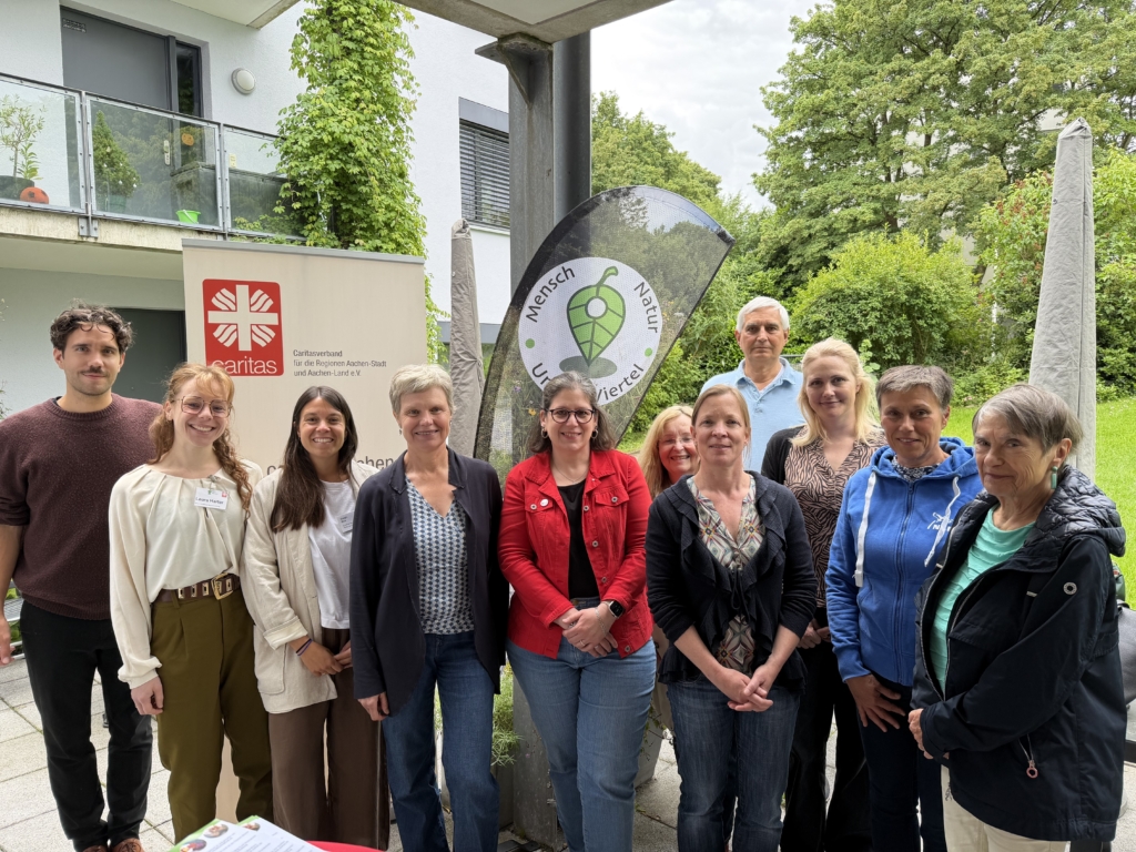 Verantwortliche der Caritas und Engagierte aus dem Viertel stehen als Gruppe zusammen, im Hintergrund ein Roll-up-Banner der Caritas und eine Beachflag mit dem Logo von Mensch - Natur - Viertel.
