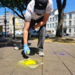 Ein Mann mit Jeans, T-Shirt und Kappe hockt auf dem Boden und sprüht bunte Schmetterlinge mit Kreidefarbe auf den Boden.