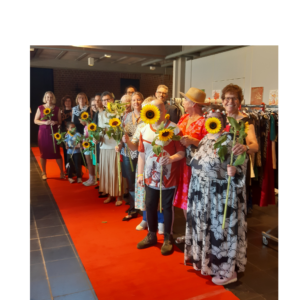 Viele Menschen stehen in bunter Secondhand-Kleidung auf einem roten Teppich. Sie halten Sonnenblumen in der Hand.