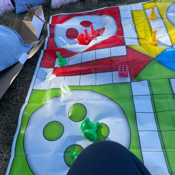 Eine übergroße Mensch-Ärgere_Dich-Nicht-Plane liegt auf der Wiese. Auf dem Spielfeld sieht man große , bunte Spielsteine. Ein Kind liegt dahinter - zu sehen sind nur die Beine und ein Stück von Arm und Hand.