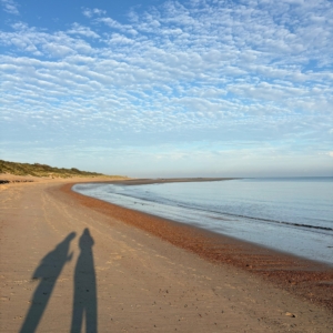 Ein Küstenabschnitt, ruhiges Wasser, Sand und Dünen am Rand. Der Schatten zweier spazierenmder Menschen ist auf dem Sand zu sehen.